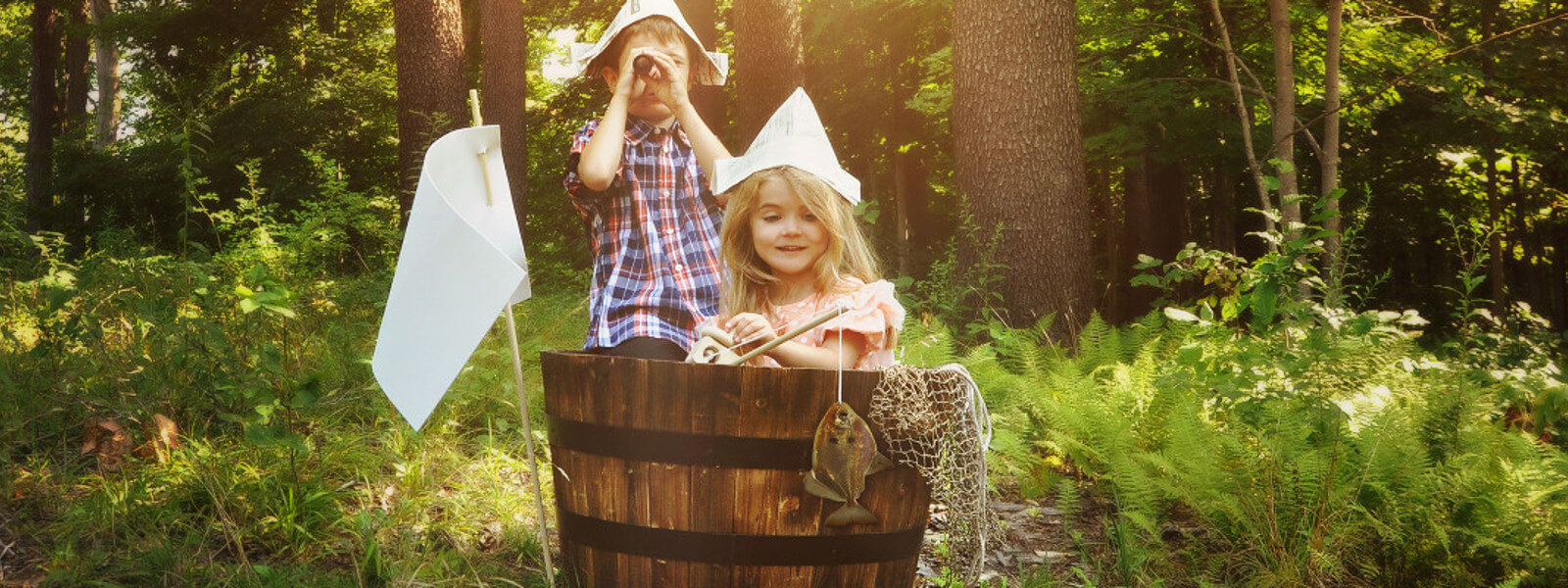 Two children sit in a wooden barrel in the woods, pretending to be sailors.