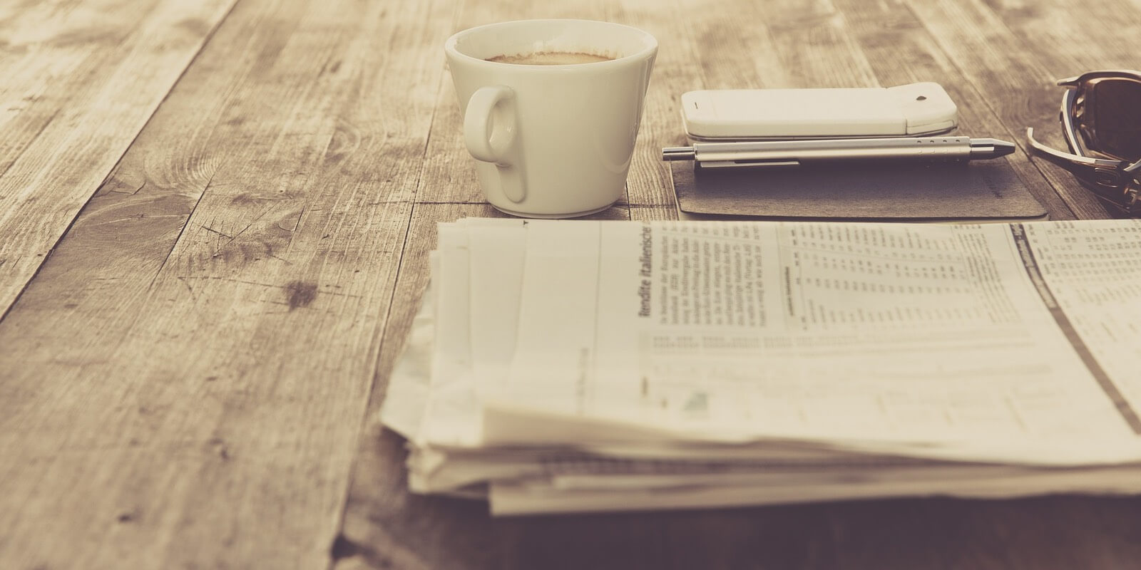 A newspaper, sits on a table next to a cup of coffee, a phone, a pen and a pair of sunglasses.