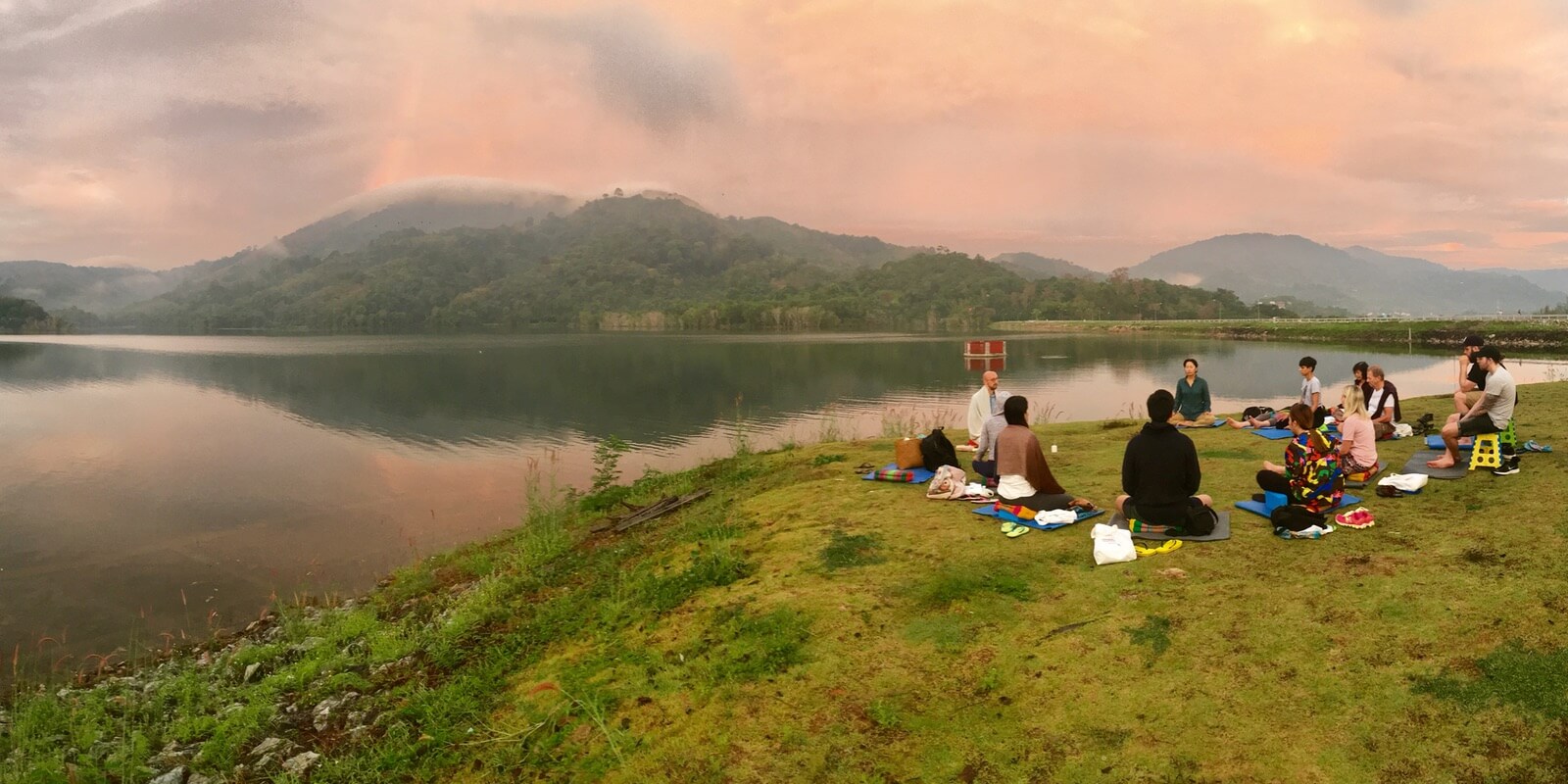 People meditate together at a lake, surrounded by tropical jungle and hills. This is the meditative lifestyle.