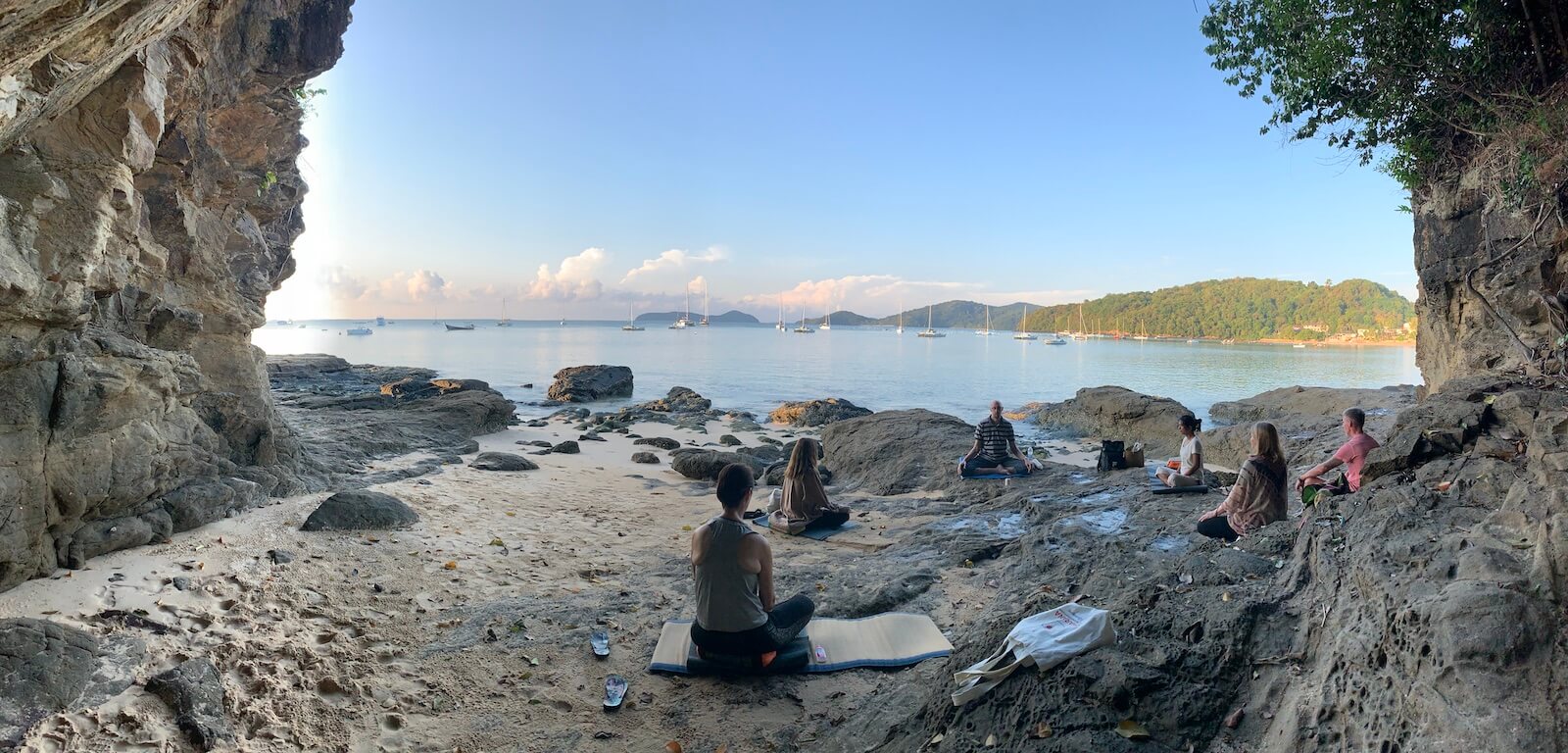 Six people are meditating in a group together at Aoyon beach in Phuket, next to a cliff.